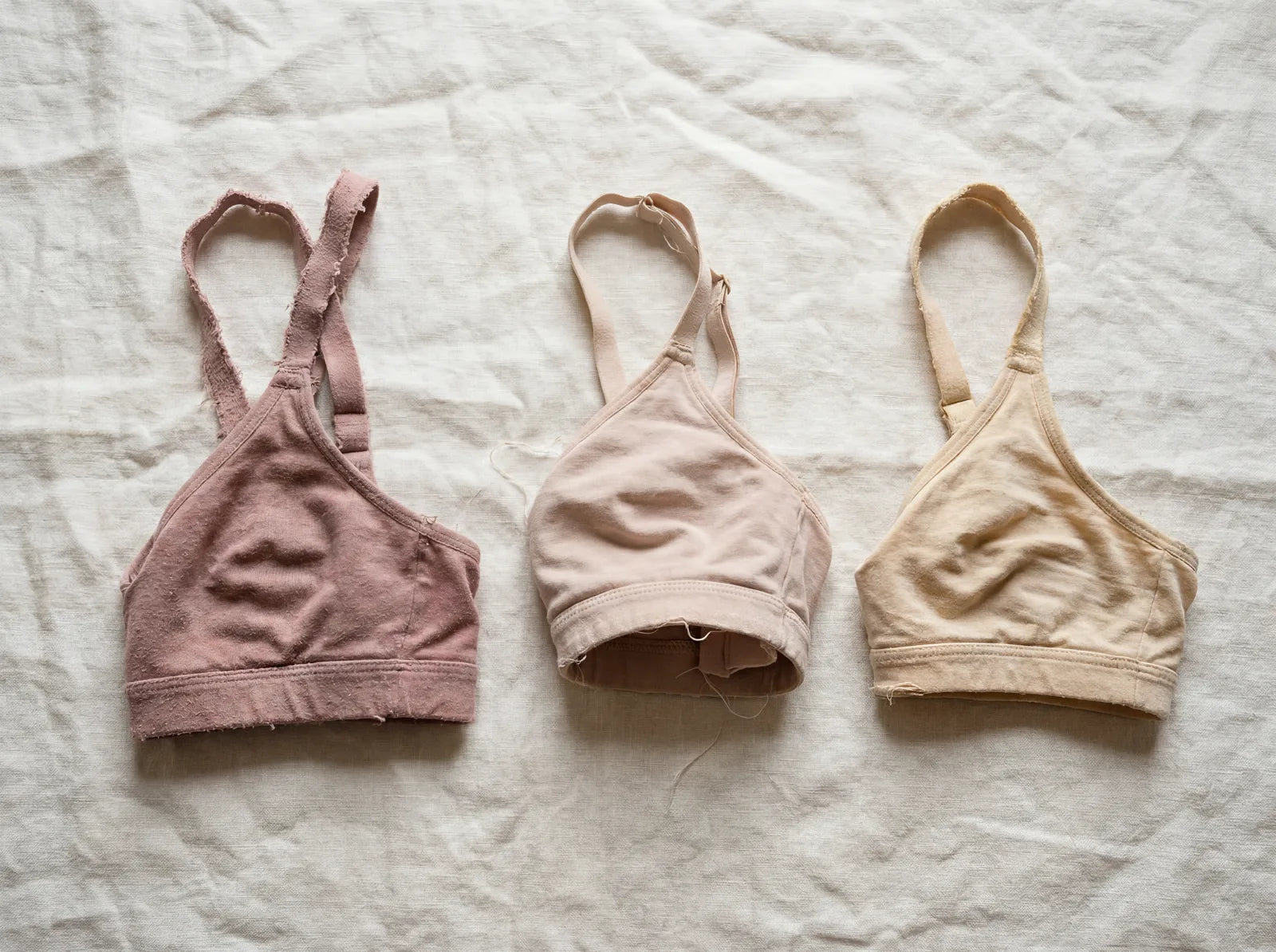 Three worn-out bras laid side by side on a rumpled linen bedspread in morning light